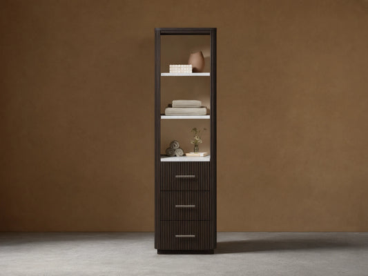 Tall dark brown Finnley bath cabinet with open shelves displaying decor/towels & three fluted drawers with silver pulls, against a beige wall.
