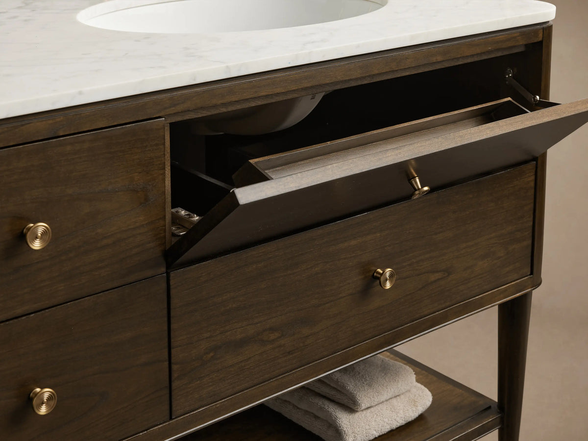 Close-up, high-angle of dark brown Laurent Double Vanity. White marble top, brass knobs. Open drawers reveal towels, perfume, comb.