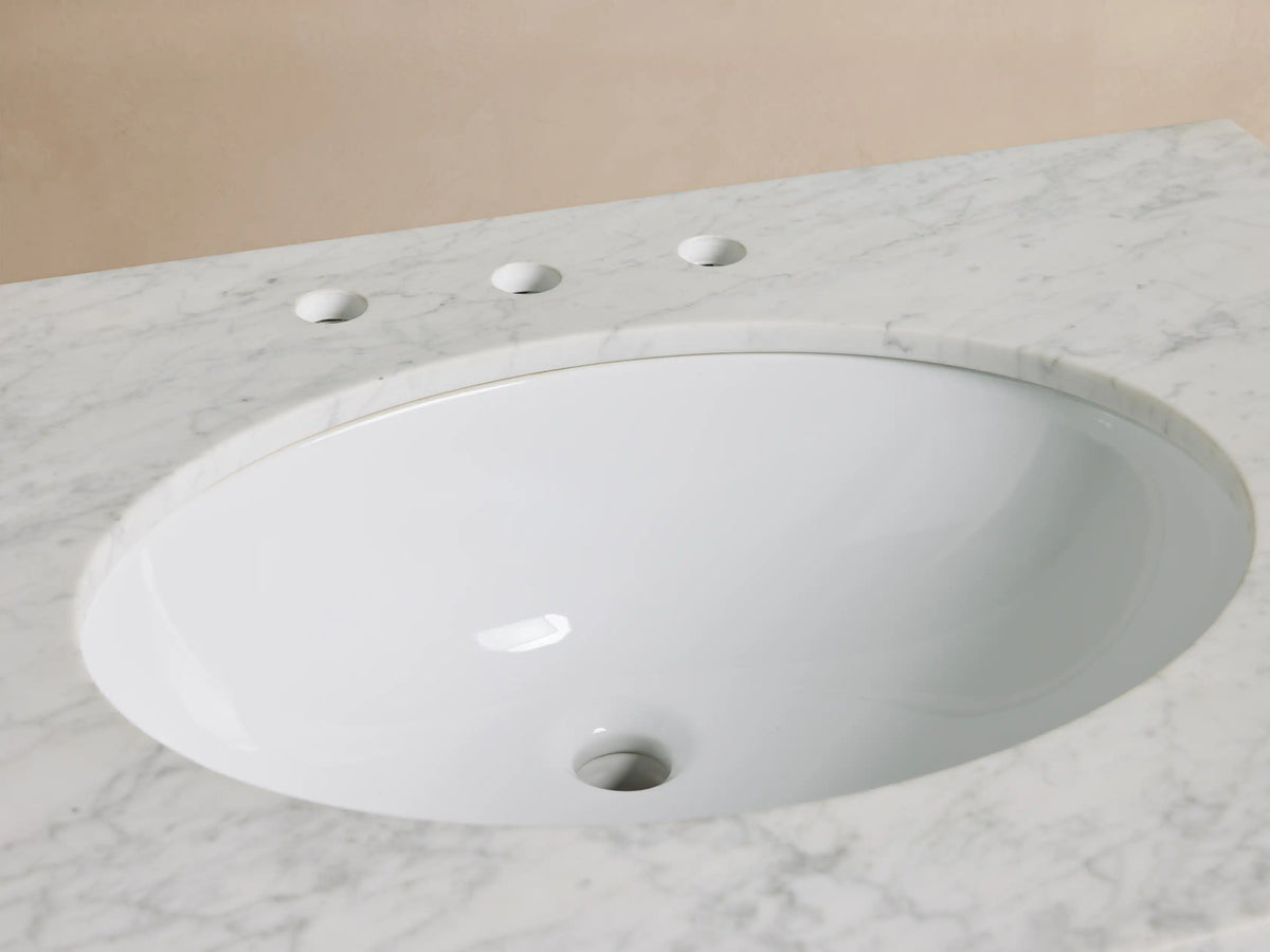 Close-up of Laurent Single Vanity: white oval sink, veined Carrara marble top, antique brass faucet, soap dispenser, candle.