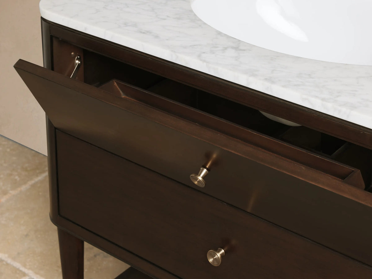 Laurent Single Vanity close-up. Dark brown wood cabinet, white marble top, gold knob. Open drawer shows beige towels, perfume.