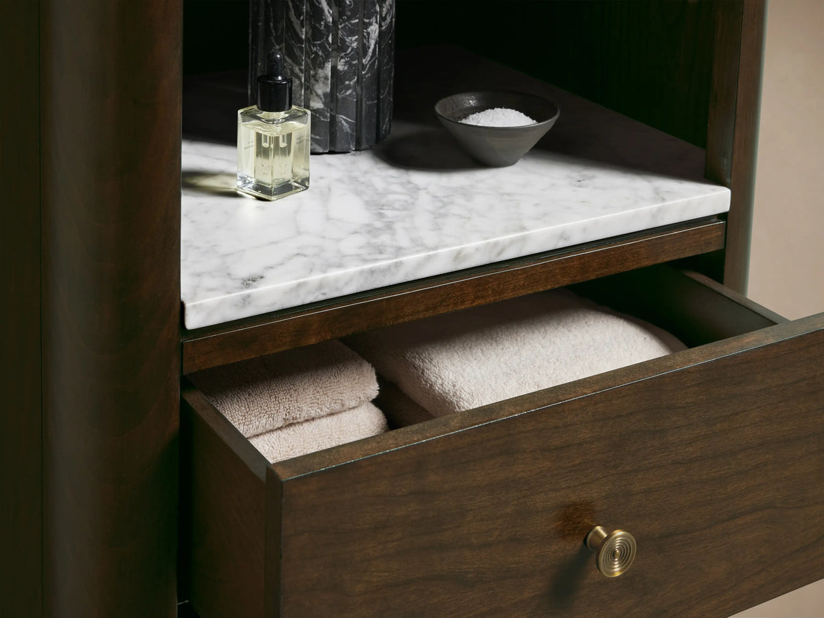 Full view of a slender Laurent Tall Bath Cabinet in Bahasa Brown wood, with open shelves, white marble top, 3 drawers with brass pulls.