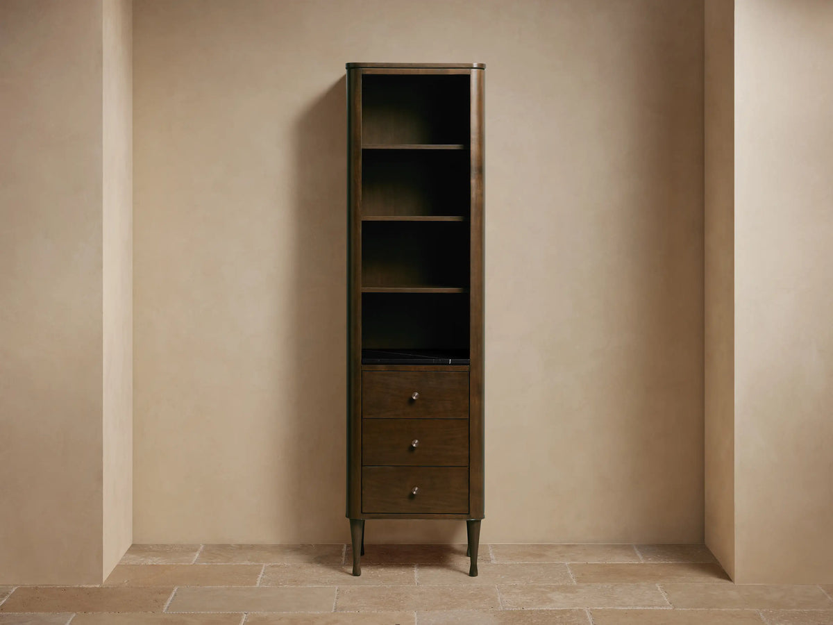 Laurent Tall Bath Cabinet: dark Bahasa Brown wood, with four open shelves holding decor and three drawers below, against a light wall.