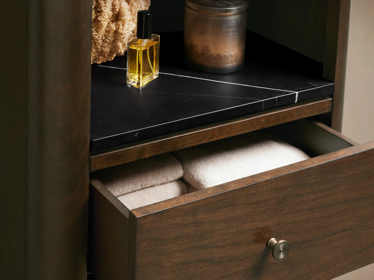 Dark brown Laurent Tall Bath Cabinet with open shelves, three drawers, and tapered legs, against a plain wall on stone floor.