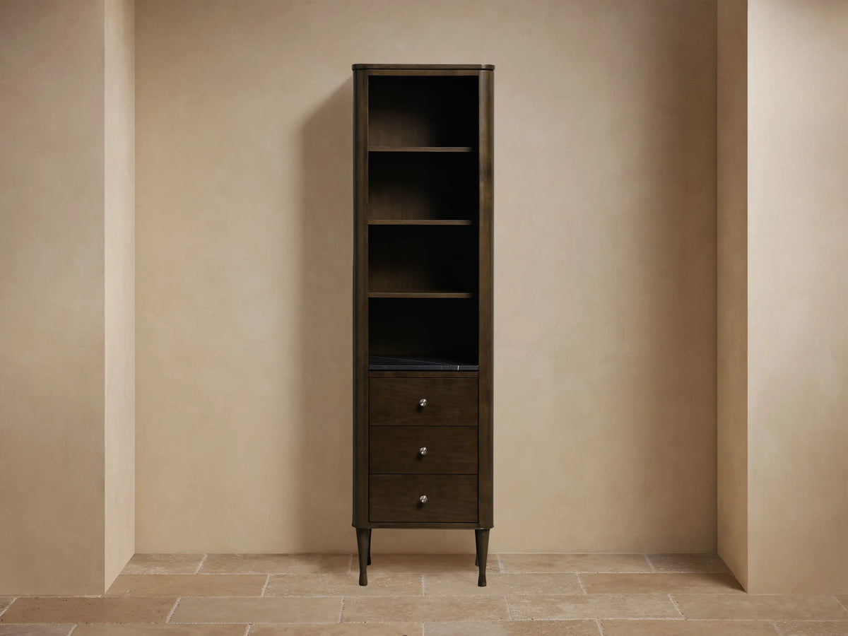 Tall Laurent Bath Cabinet, dark Bahasa Brown finish, with open shelves holding decor & towels, above three drawers on a stone floor.