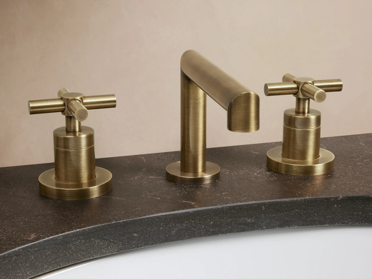 Lomas Faucet in antique brass with cross-styled handles on a marble countertop, with an oval sink and matching drain.