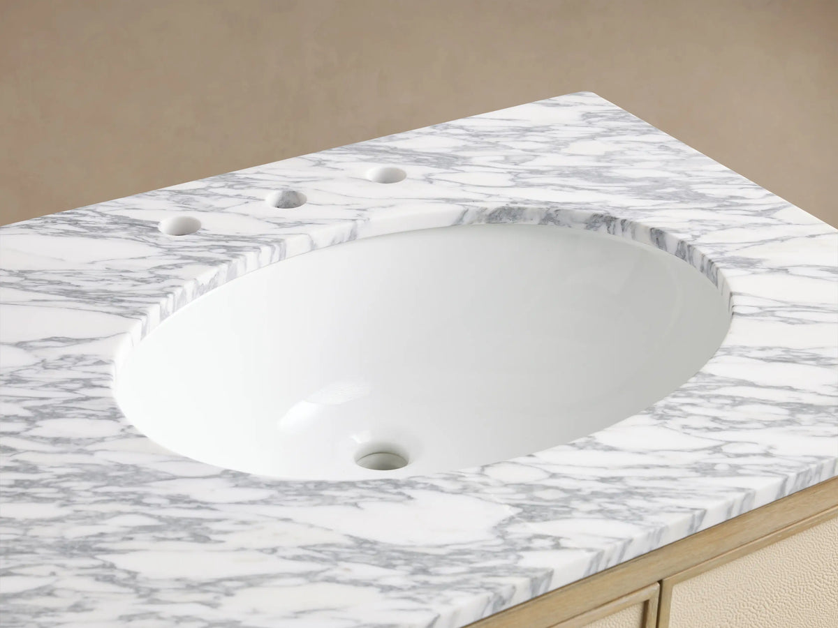 Close-up of Malone Vanity's white Carrara marble top with grey veins, oval undermount sink, brass faucet, and bone oak vanity base.