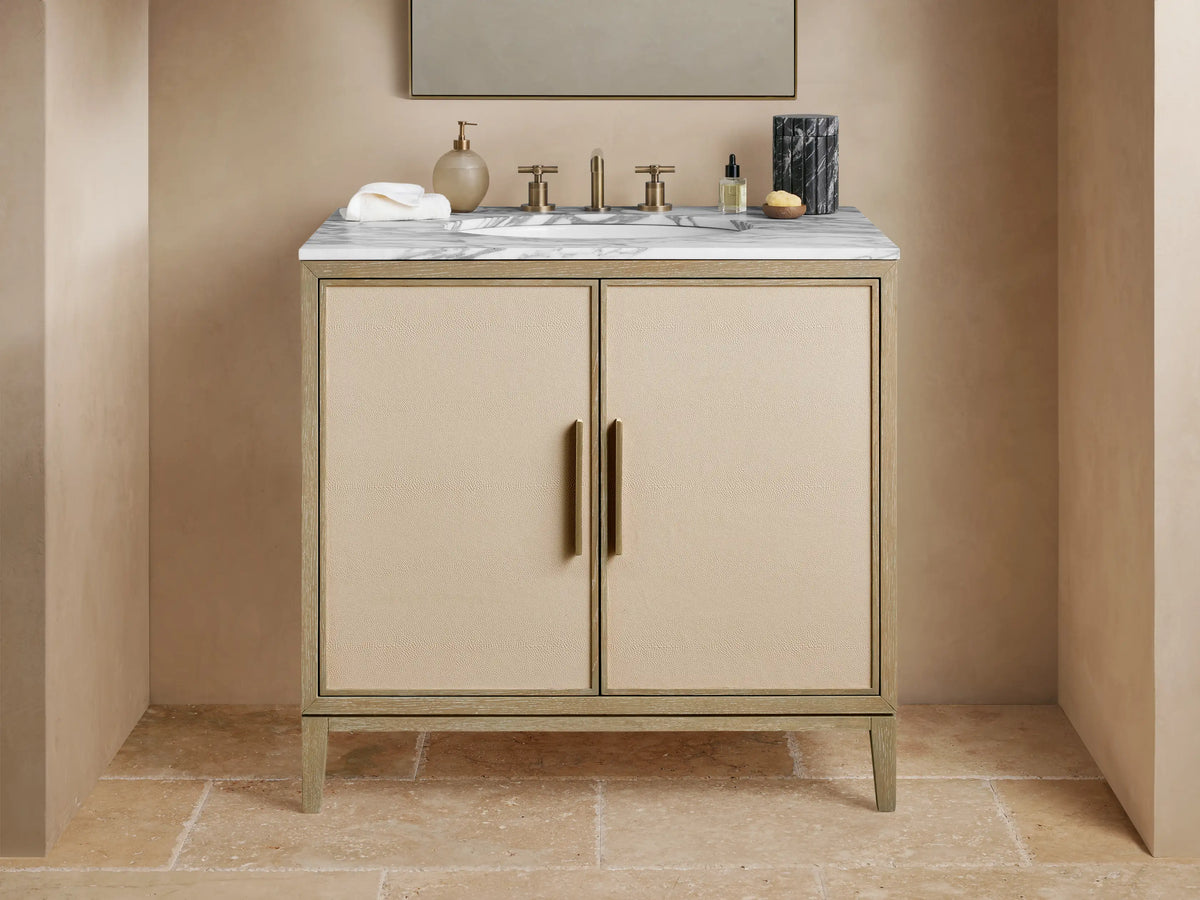 Malone Single Vanity with light wood frame, beige shagreen doors, white marble top, and brass hardware, in a bathroom setting.