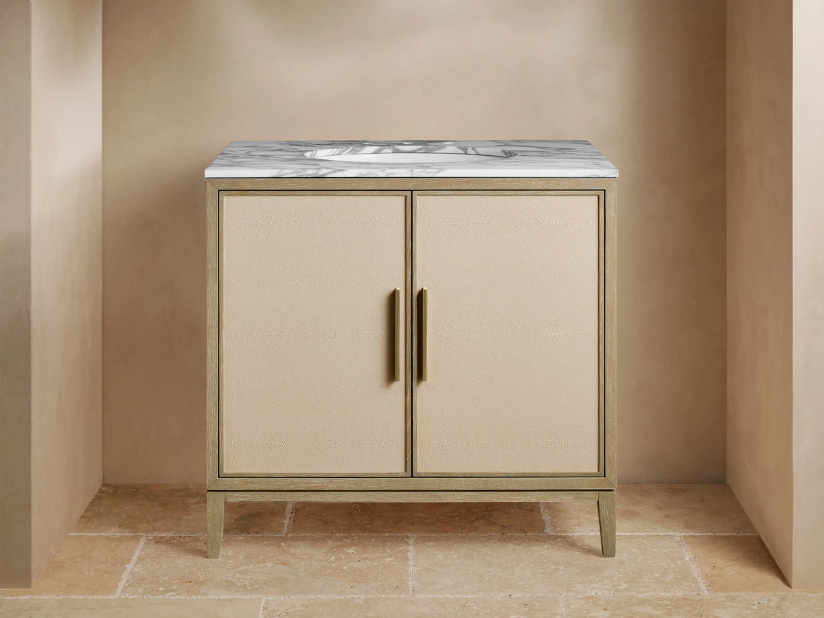 Malone Single Vanity with light wood frame, beige shagreen doors, white marble top, and brass hardware, in a bathroom setting.