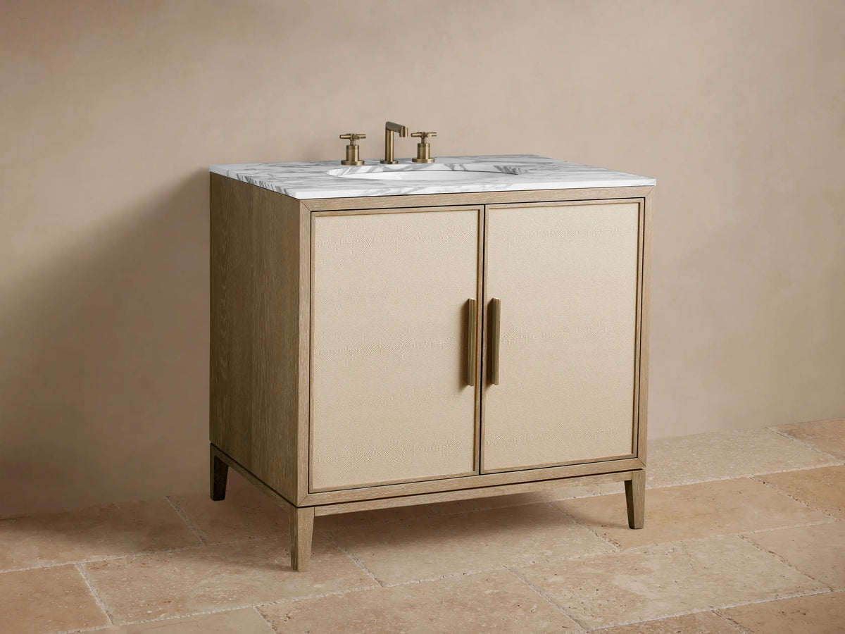 Close-up of Malone Single Vanity with open beige textured door, antique brass handle, revealing white towels, soaps, perfume inside.