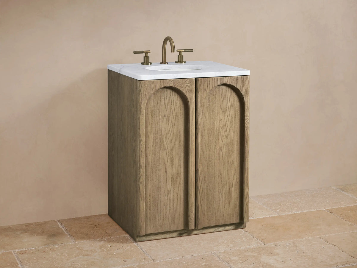 Provo Powder Vanity with oak wood finish, featuring dual arched doors, a white stone top, and brass faucets, set against a neutral-toned wall.