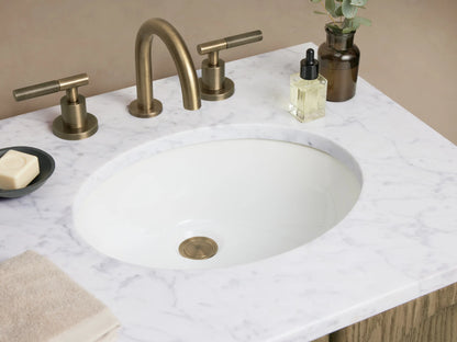 Oak wood vanity with two arched soft-close doors, white stone top, and dual brass faucets. Beige wall and tiled floor.