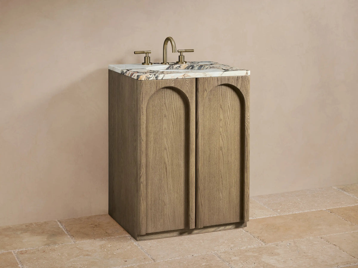 Provo Powder Vanity with oak wood finish, honed marble top, brass faucet, oval undermount sink, two arched doors, set against a neutral-toned bathroom.
