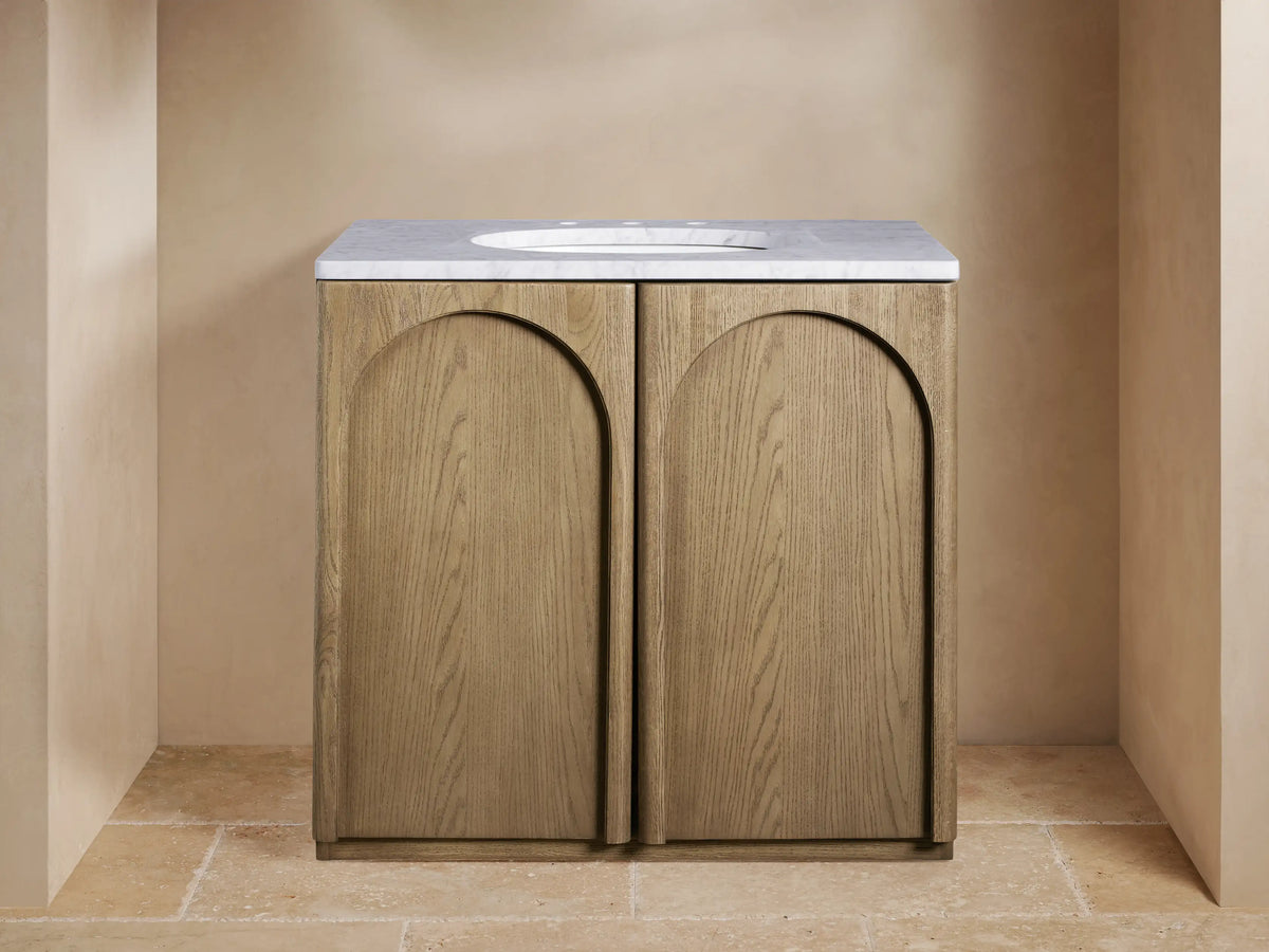 Provo Single Vanity: Light oak wood with visible grain, arched doors, white marble top, oval sink, and brass faucet in a modern setting.