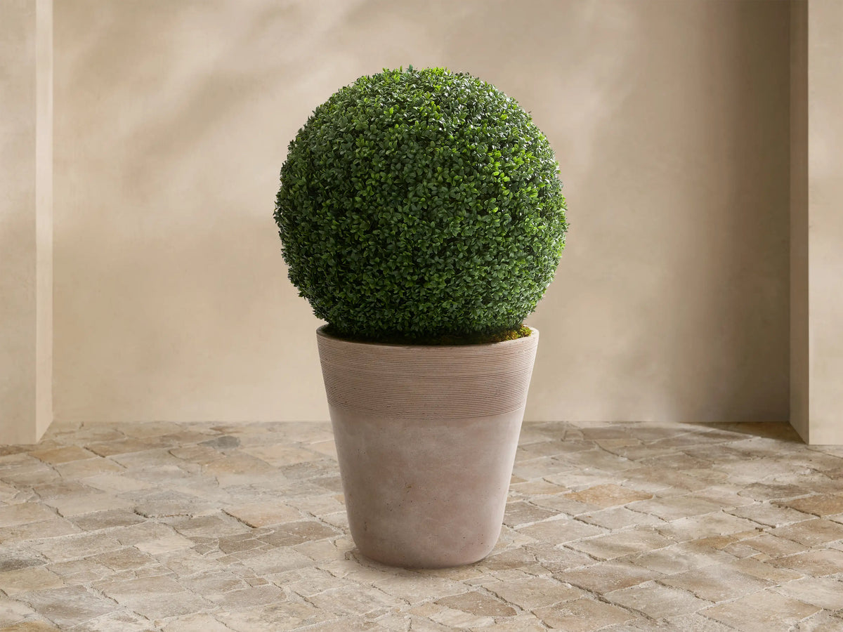 Spherical green faux boxwood in a light beige, ribbed, conical planter, on a stone floor against a tan wall.
