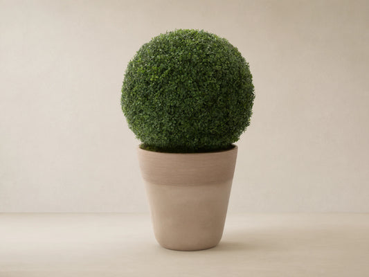Spherical green faux boxwood in a light beige, ribbed, conical planter, on a stone floor against a tan wall.