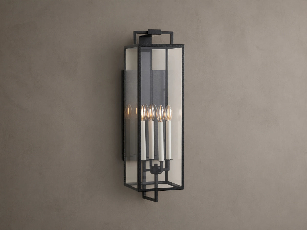 Hendricks Outdoor Sconce: black iron frame with clear glass panels, four lit candle-style bulbs, mounted vertically on a taupe wall.