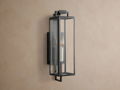 Hendricks Outdoor Sconce: dark iron rectangular lantern with clear glass panels, glowing candle-style bulb, mounted on a brown wall.