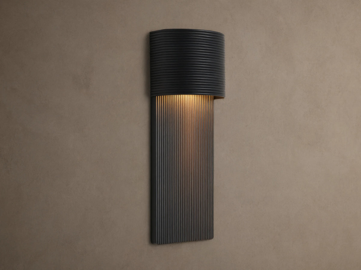 Fultz Outdoor Sconce, a soft black wall-mounted fixture with a horizontally ridged top and vertically ridged base, glowing warm light.