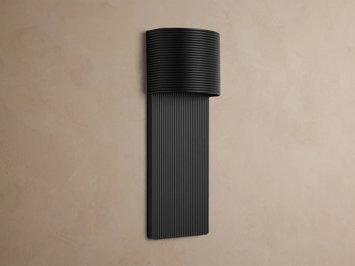 Fultz Outdoor Sconce, a soft black wall-mounted fixture with a horizontally ridged top and vertically ridged base, glowing warm light.