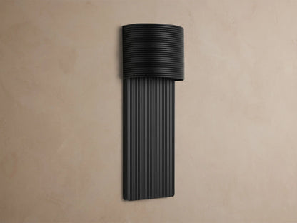 Fultz Outdoor Sconce, a soft black wall-mounted fixture with a horizontally ridged top and vertically ridged base, glowing warm light.