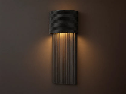 Fultz Outdoor Sconce, a soft black wall-mounted fixture with a horizontally ridged top and vertically ridged base, glowing warm light.
