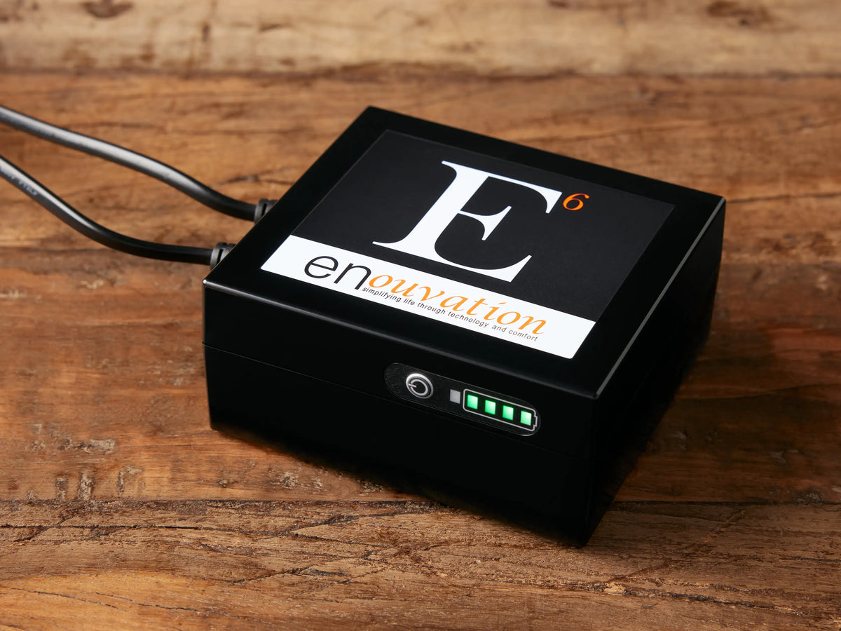 Black rectangular Rowland Collection Battery Pack with Enouvation E6 logo, two cables, and full green LED power indicator, on wood.