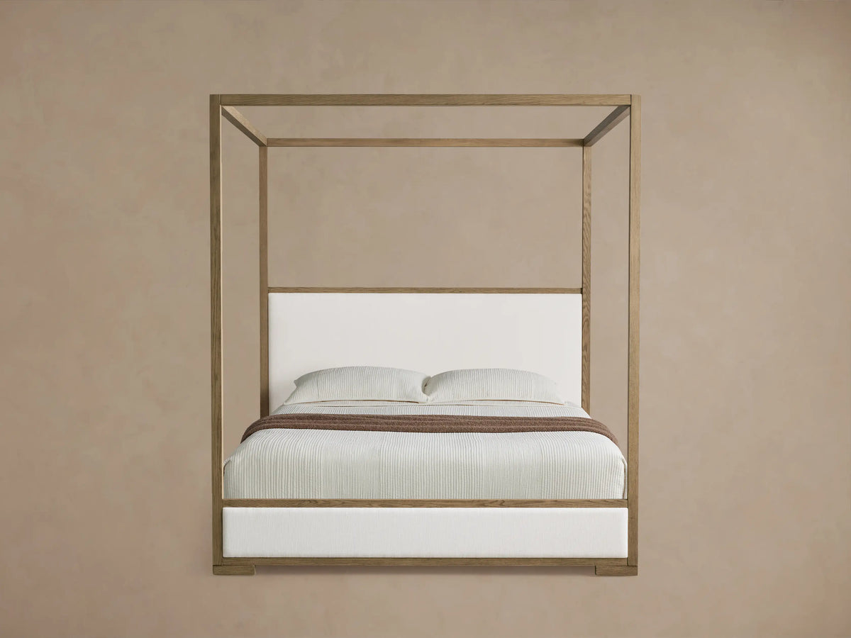 Darien Canopy Bed: light oak wood frame, white upholstered headboard & base, light bedding, brown throw, angled view.