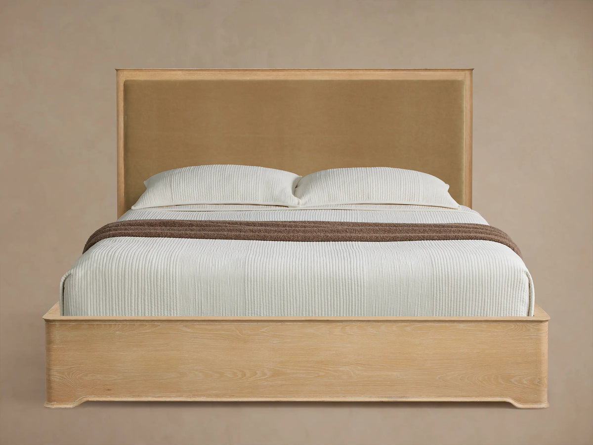Light oak Conley Panel Bed with tan fabric headboard, white bedding, and brown throw, angled on a wooden floor.