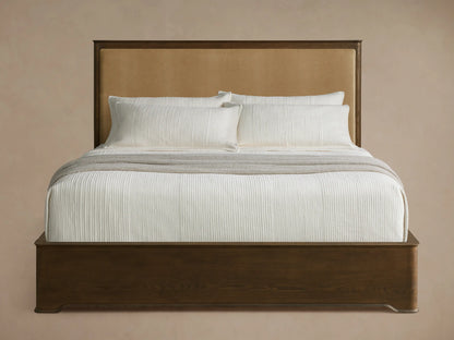 Conley Panel Bed with dark wood frame and tan upholstered headboard, white bedding, and grey throw blanket.