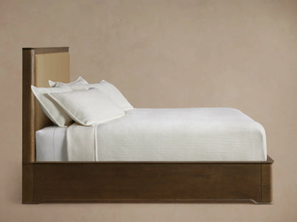 Close-up of Conley Panel Bed headboard. Dark oak frame with visible wood grain, curved top edge, and light tan fabric panel.