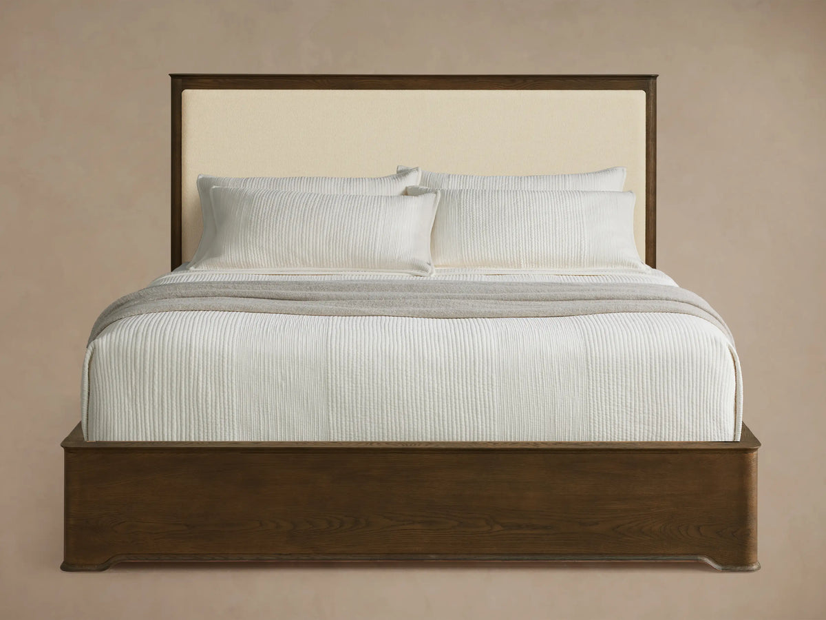 Conley Panel Bed with dark oak frame & cream upholstered headboard. Bed has white textured bedding & a gray throw blanket.