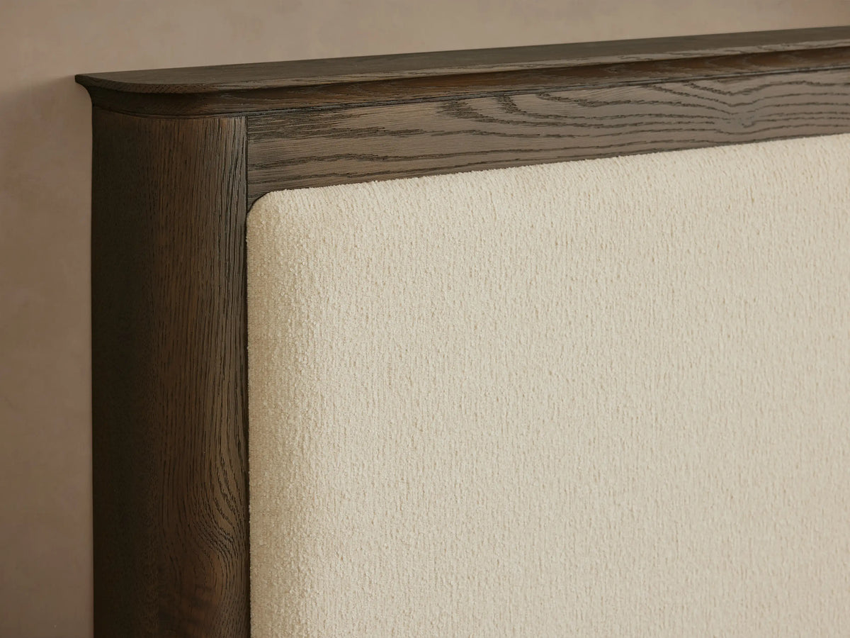 Conley Panel Bed: Dark oak wood frame with a cream upholstered headboard, white quilted bedding, and a grey throw.