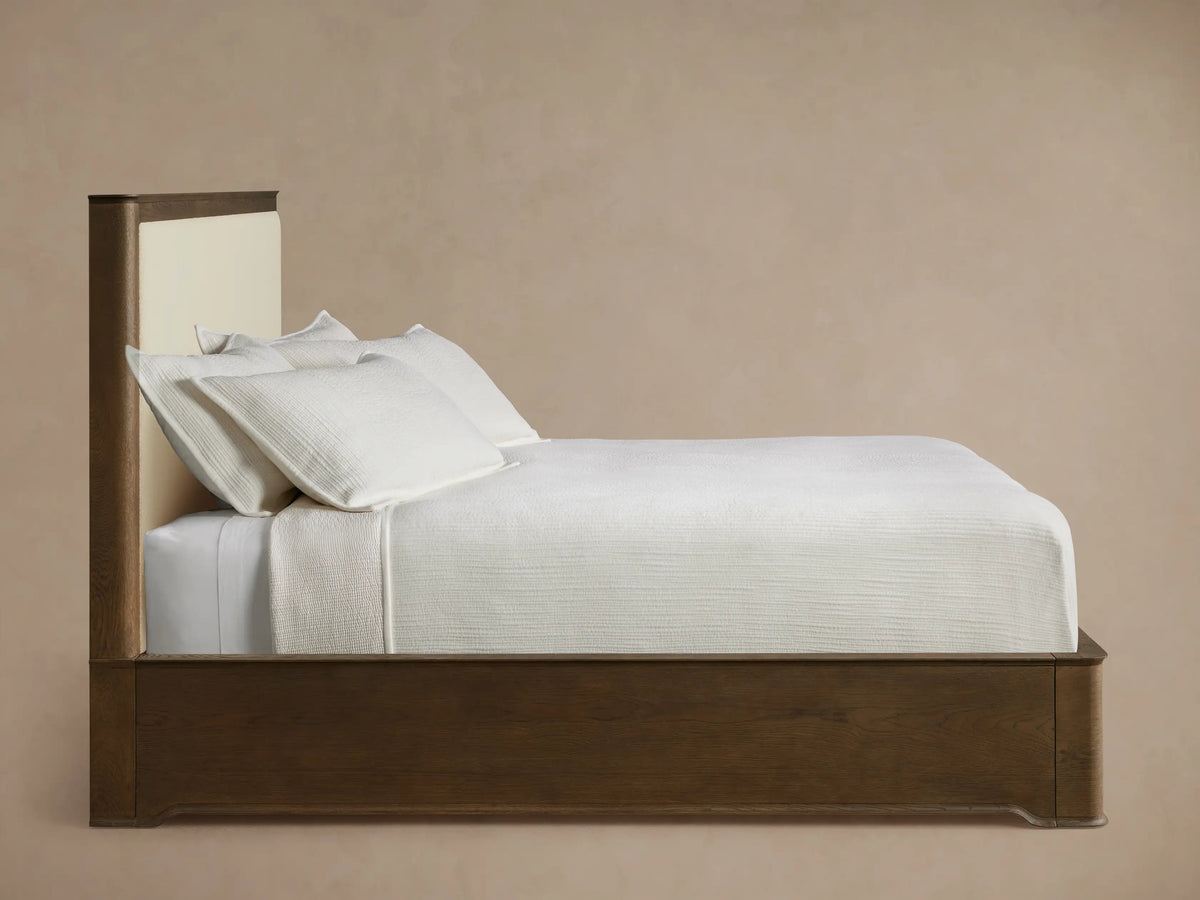 Conley Panel Bed headboard. Dark oak frame with curved top and visible grain, bordering a light cream upholstered fabric panel.