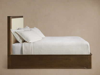 Conley Panel Bed headboard. Dark oak frame with curved top and visible grain, bordering a light cream upholstered fabric panel.