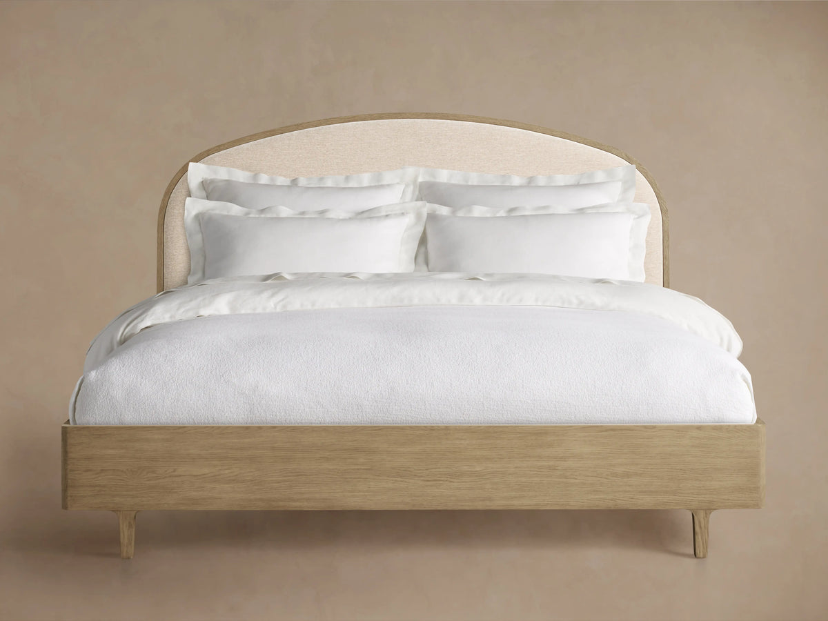 Full shot of a light oak Danny Bed with an arched beige upholstered headboard and clean white bedding against a tan wall.