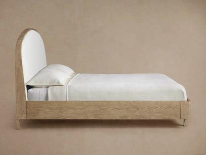 Close-up of Danny Bed headboard. Curved light oak wood frame with visible grain bordering light beige textured upholstered fabric.