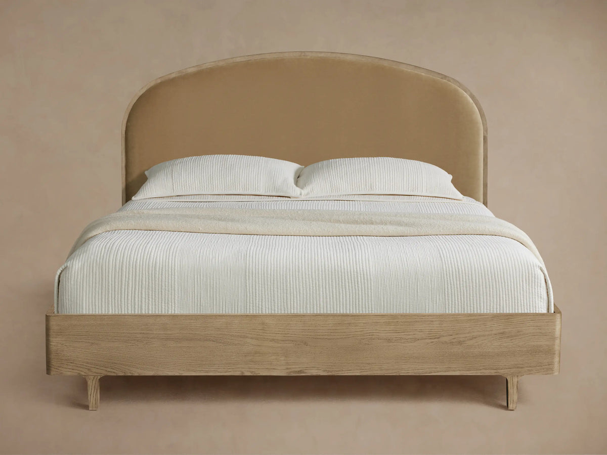 Danny Bed with curved tan upholstered headboard & light oak frame, white corrugated bedding & throw blanket on dark wood floor.