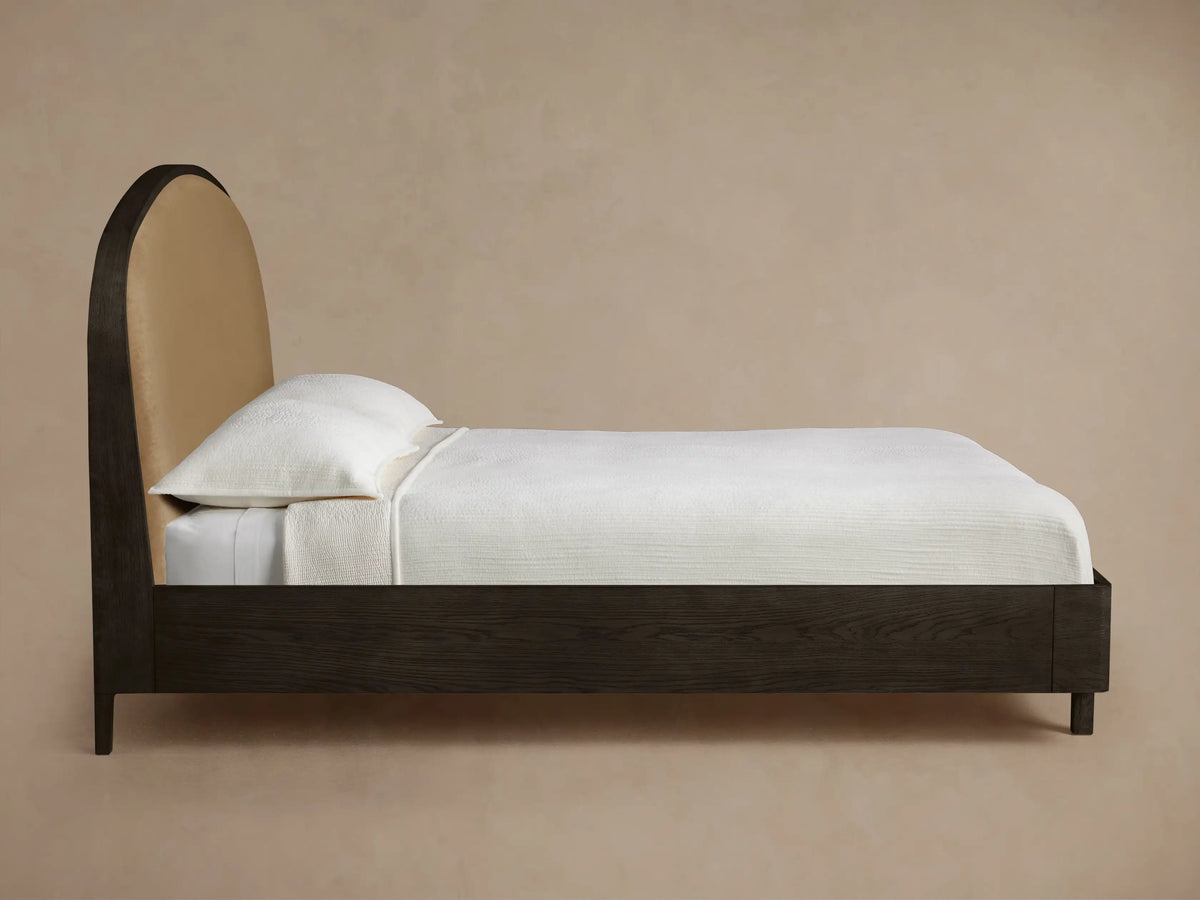 Danny Bed headboard. Close-up of curved dark oak frame with visible grain, bordering soft, light brown upholstered fabric.