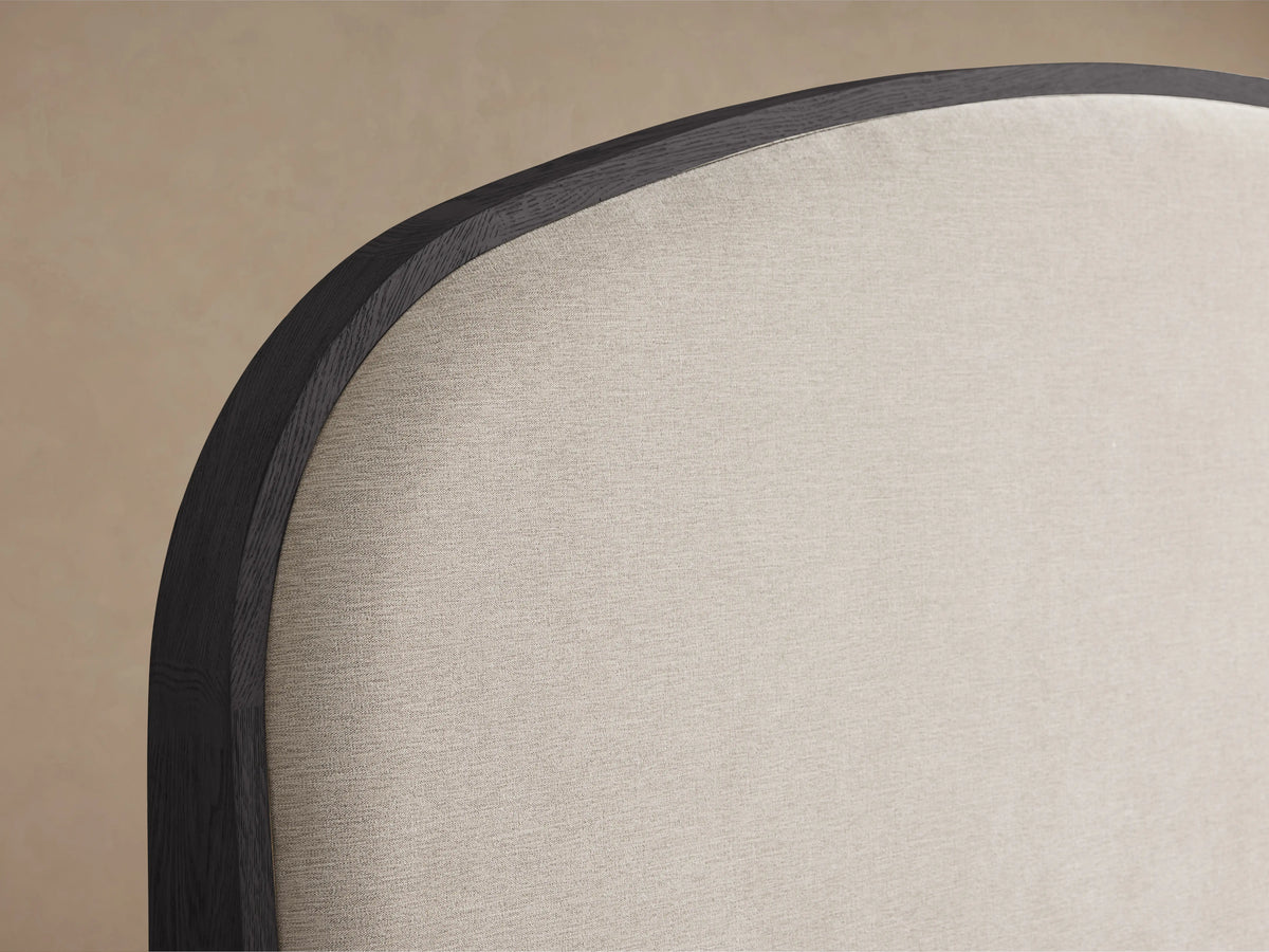 Close-up of Danny Bed headboard. Curved dark oak frame outlining light beige textured fabric. Against a neutral tan wall.