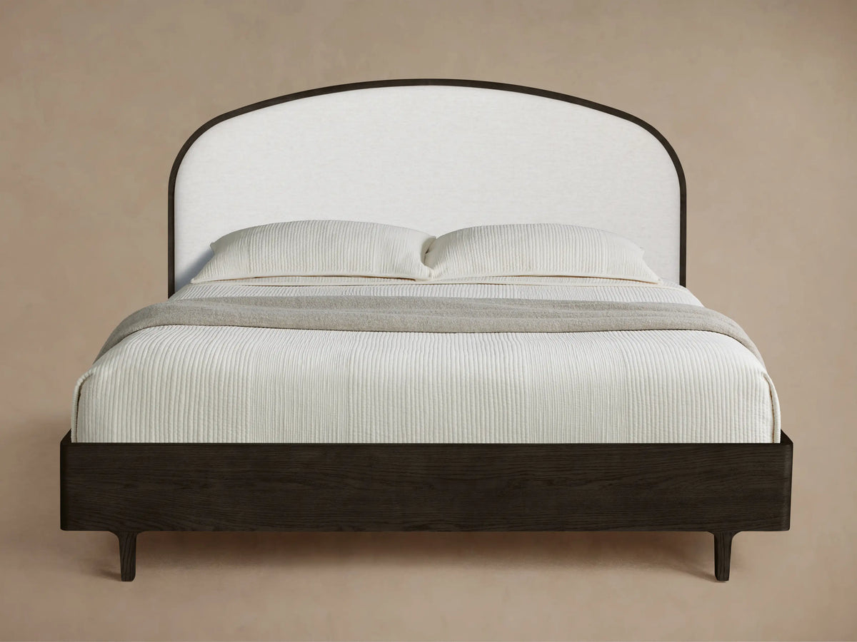 Danny Bed with dark wood frame, light upholstered curved headboard, white textured bedding, and a gray throw on wooden floor.