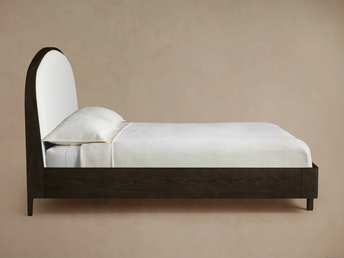 Close-up of Danny Bed headboard: curved dark oak wood frame outlining light textured fabric upholstery.