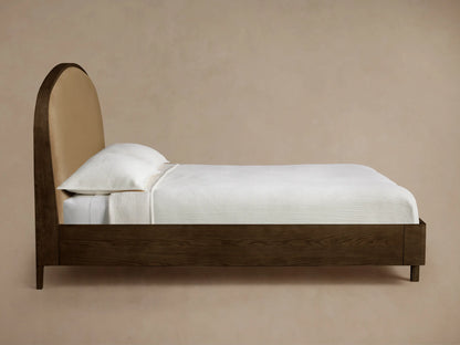 Side profile of the Danny Bed with an arched tan upholstered headboard and dark brown wooden frame, made with white bedding.