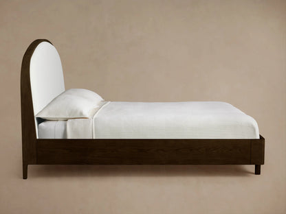 Side view of the Danny Bed, featuring a dark brown oak frame and an arched white upholstered headboard, dressed in white bedding.