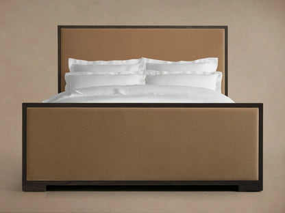Darien Panel Bed with high footboard, oak frame with brown upholstered panels, white bedding against beige wall.