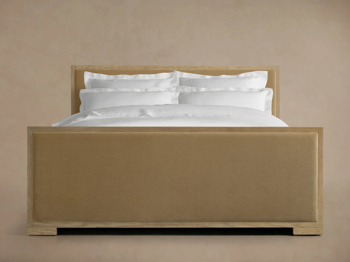 Darien Panel Bed with high footboard, oak frame, brown upholstered panels, white bedding, against a beige background.