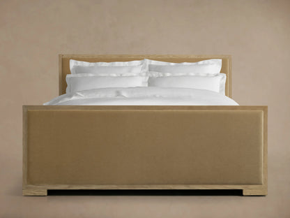 Darien Panel Bed with high footboard, oak frame, brown upholstered panels, white bedding, against a beige background.
