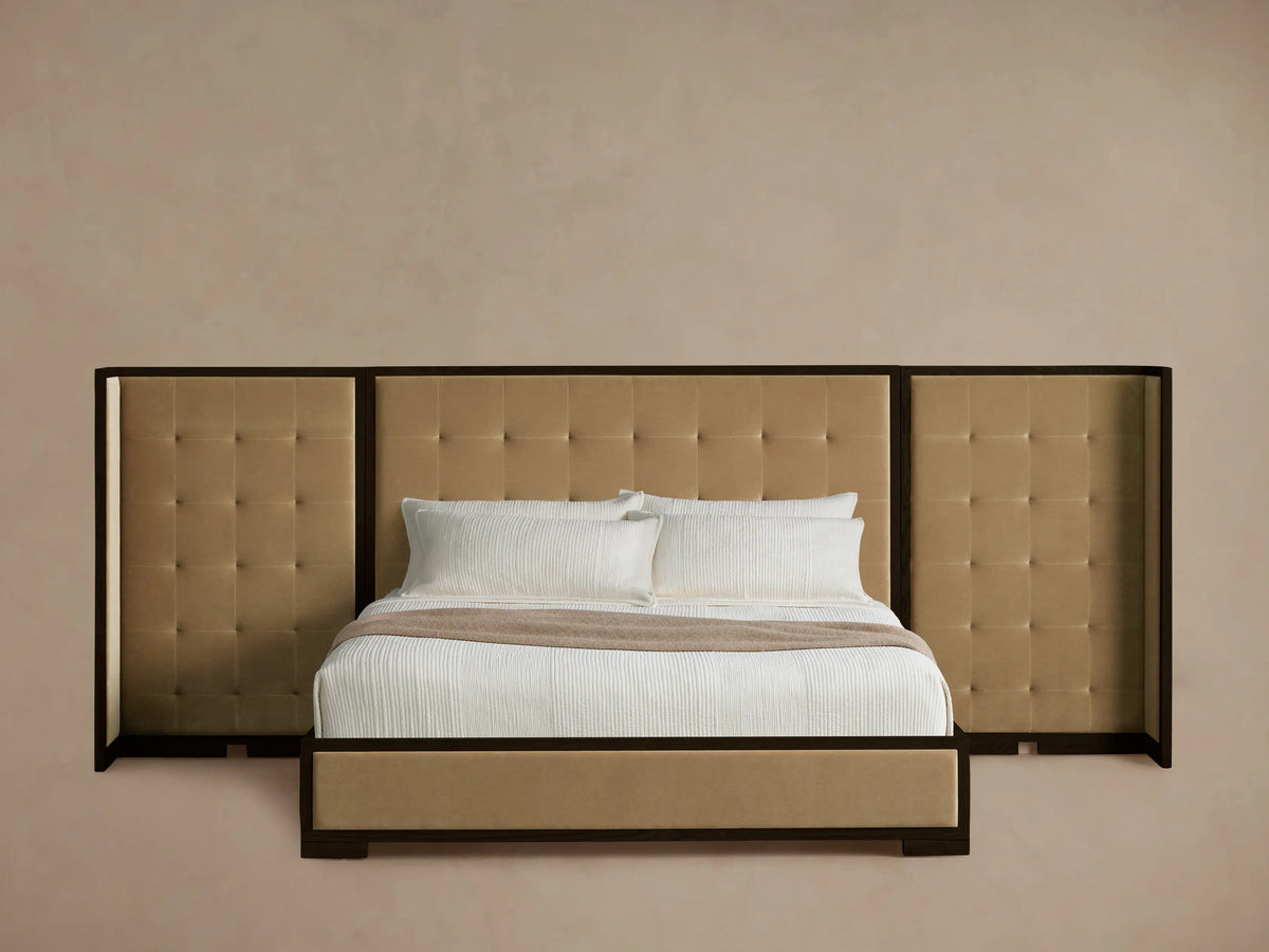 Darien Extended Box-Tufted Shelter Bed in tan upholstery with dark wood frame, white bedding & brown throw on wood floor.