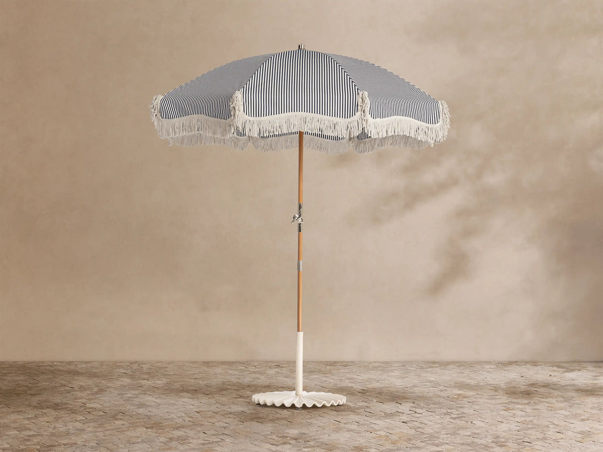 Front view of a navy and white striped outdoor umbrella with white fringe, a wooden pole, and a white scalloped base.