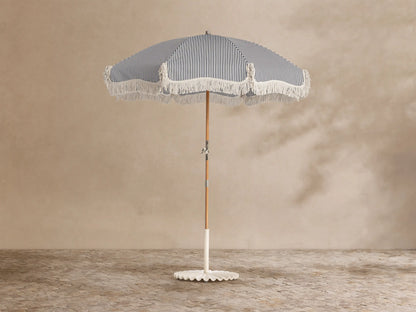 Front view of a navy and white striped outdoor umbrella with white fringe, a wooden pole, and a white scalloped base.