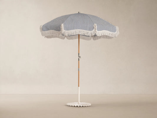 Front view of a navy and white striped outdoor umbrella with white fringe, a wooden pole, and a white scalloped base.